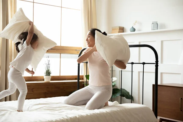 Happy Asian mom and little daughter engaged in pillow fight