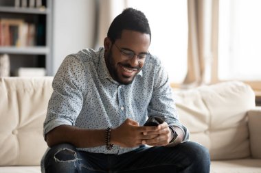 Gülümseyen Afrikalı adam kanepeye oturup akıllı telefondan mesaj atıyor.