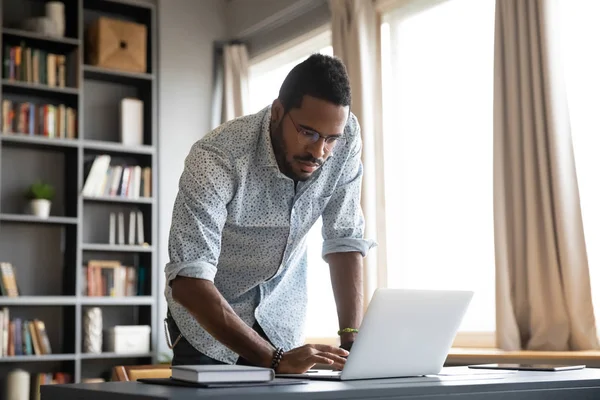 Fokuserad afrikansk affärsman står vid hemmakontoret skrivbord med laptop — Stockfoto