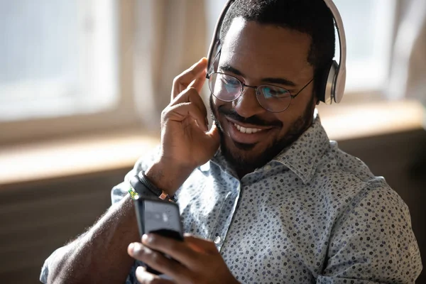 Sorrindo homem africano usando fones de ouvido ouvir música móvel no smartphone — Fotografia de Stock