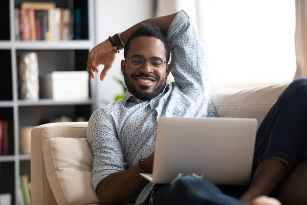 Χαμογελώντας νεαρός ενήλικας Αφρικανός χρησιμοποιώντας φορητό υπολογιστή χαλαρώνοντας στον καναπέ — Φωτογραφία Αρχείου