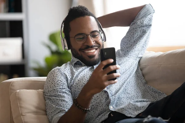 Glad afrikansk man i hörlurar lyssnar mobilmusik på soffan — Stockfoto