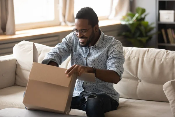 Gülümseyen Afrikalı müşteri kanepenin üzerinde karton kutu paketi açıyor. — Stok fotoğraf