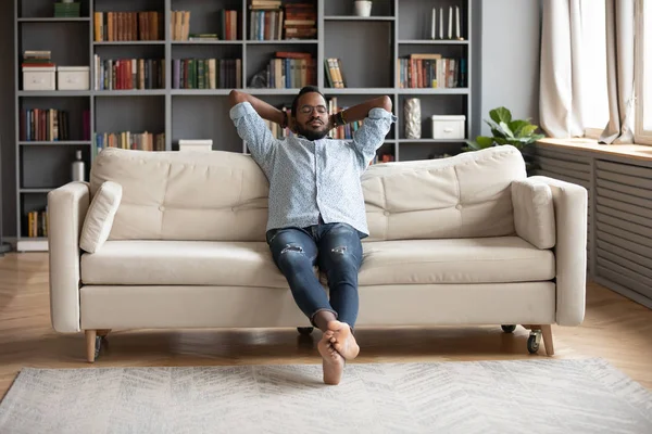 Gelassener barfüßiger afrikanischer Mann, der sich auf Sofa-Händen hinter dem Kopf ausruht — Stockfoto