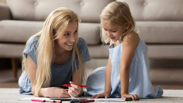 Joven madre acostada en la alfombra, enseñando fotos para colorear hija pequeña . — Foto de Stock