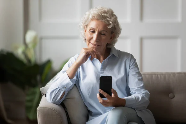 Happy older woman sitting on comfortable couch, looking at smartphone screen. — Stock Photo, Image
