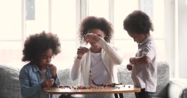 Caring african mommy helping two children stringing necklace of beads