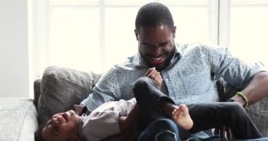 Funny happy african daddy tickling cute small son on sofa