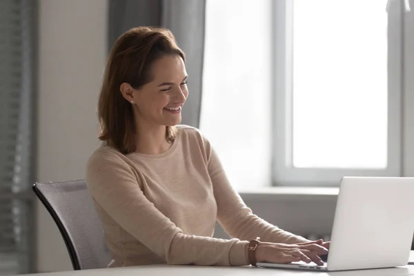 Feliz mujer milenaria sonriendo trabajando en el ordenador portátil en el lugar de trabajo — Foto de Stock