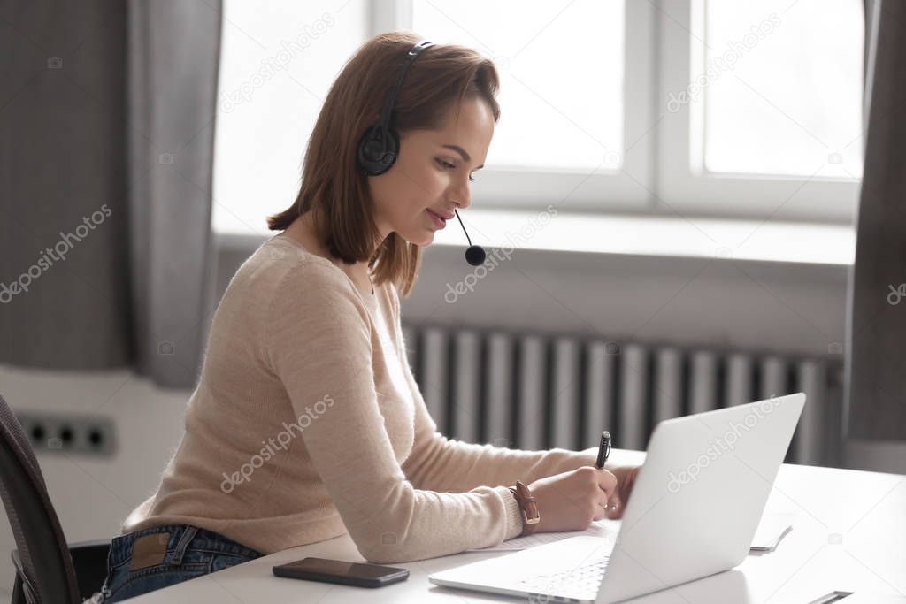 Focused female in headset make notes watching webinar on laptop