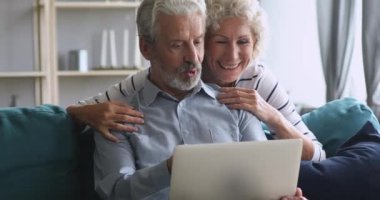 Happy elderly woman peeking from husband shoulder, looking at screen.