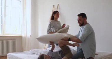 Happy young parents beating lying on bed little children with pillows.