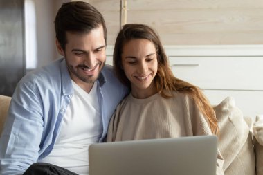Smiling millennial couple relax on couch using laptop