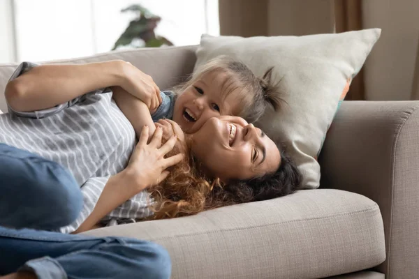 Juguetona hija preescolar tumbada en un sofá con una madre sonriente . — Foto de Stock