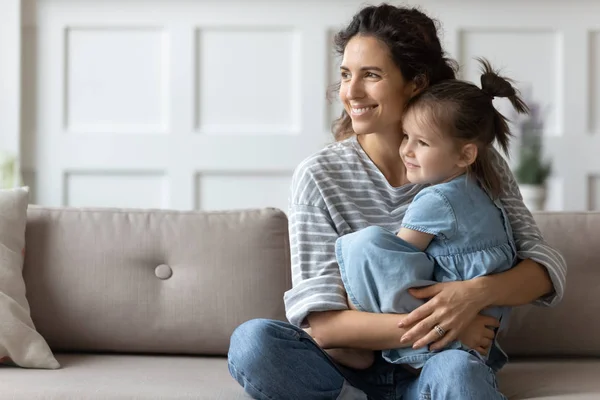 Soñadora cariñosa madre rizada abrazando pequeña hija preescolar, mirando hacia otro lado . —  Fotos de Stock