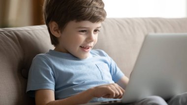 Small kid boy resting on couch, using educational app on laptop.