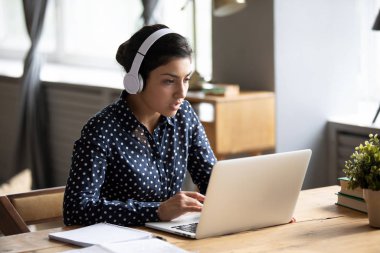 Focused Indian female in headphones watch webinar on laptop
