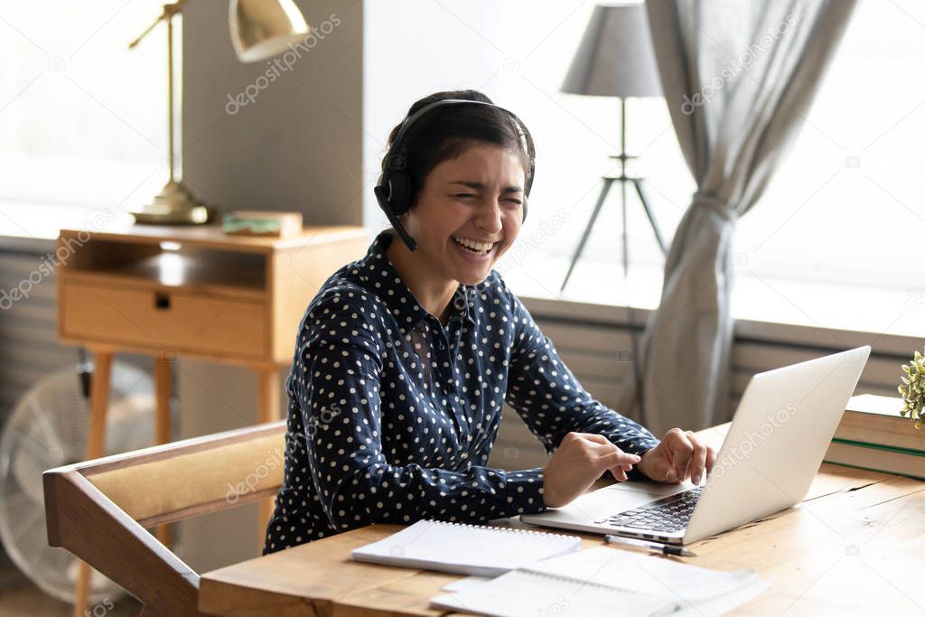 Overjoyed Indian girl in headphones laugh using laptop at home