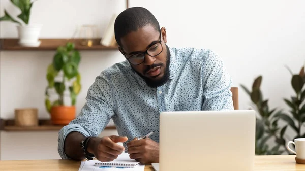 Сосредоточенный афроамериканский бизнесмен, использующий ноутбук, пишет заметки — стоковое фото