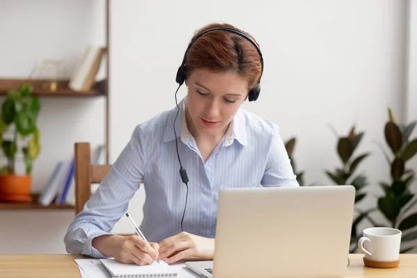 Focused woman in headphones using laptop, writing notes — 图库照片