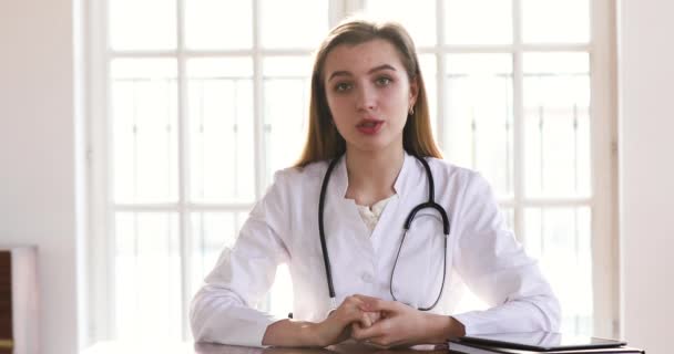 Молода жінка-лікар носить білу форму, дивлячись на розмову з камерою — стокове відео