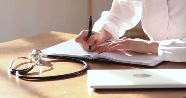 Жінка-лікар одягнена в білу уніформу робить нотатки в блокноті, крупним планом — стокове відео