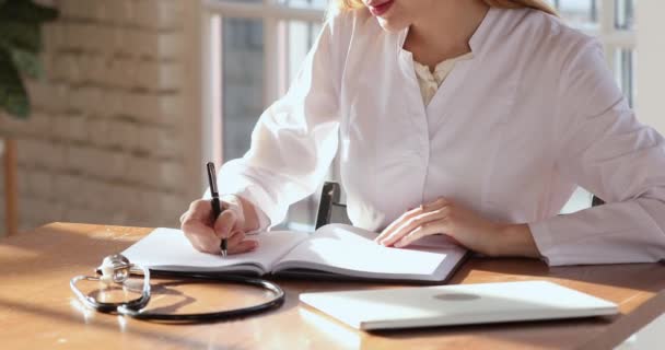 Молода жінка-лікар пише нотатки в блокноті сидіти за столом — стокове відео