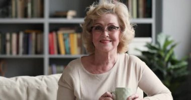 Happy older woman holding tea cup sitting on sofa, portrait
