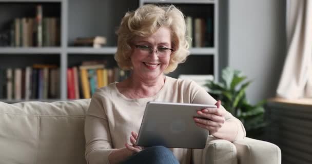 Happy mature woman using digital tablet computer sitting on couch — Stockvideo