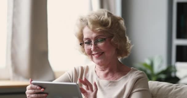 Усміхнена старша леді носить оптичні окуляри, використовуючи планшет вдома — стокове відео