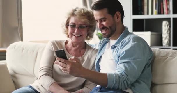 Hijo joven mostrando fotos divertidas usando teléfono inteligente con mamá madura — Vídeo de stock