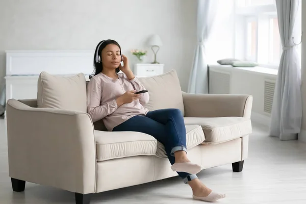 Biracial woman relax on couch listen to music using cell