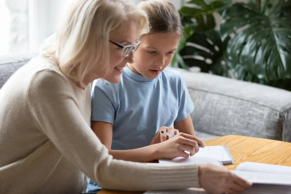 Happy older woman teacher helping school girl with home tasks.