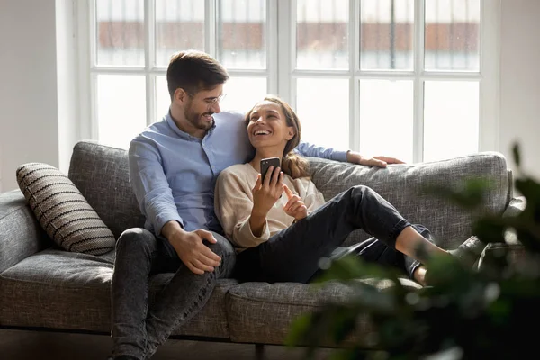 Millennial couple in love seated on couch talking using smartphone — Stock Photo, Image
