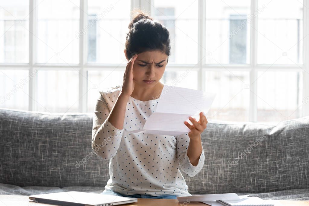 Frustrated indian woman sitting indoors holding letter reading bad news