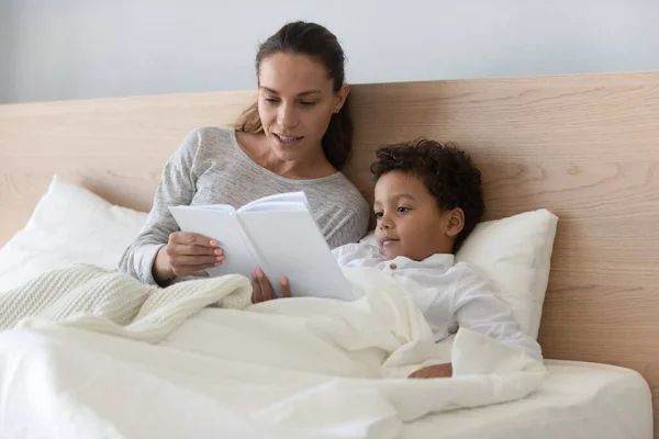 Loving mother reading book to African American son in bed