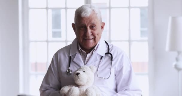 Un vieux pédiatre souriant tient un ours en peluche en regardant une caméra — Video