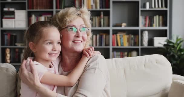 Улыбающаяся старшая бабушка обнимает защищать симпатичную внучку глядя в сторону мечтая — стоковое видео