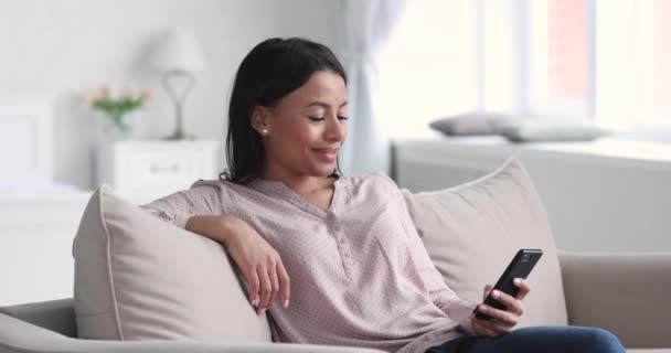 Sorrindo relaxado Africano jovem mulher usando telefone inteligente no sofá — Vídeo de Stock