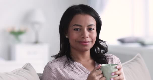 Sonriente mujer africana beber té mirando a la cámara en el sofá — Vídeos de Stock