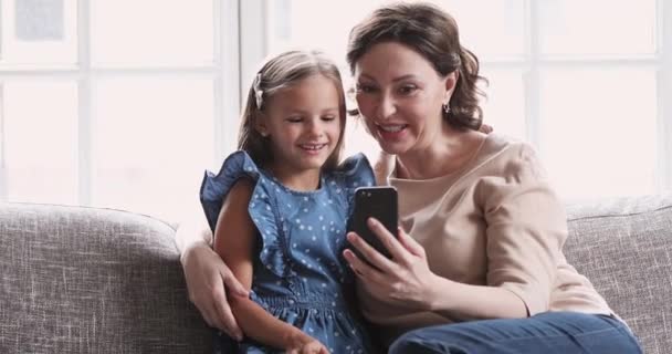 Mature grandma hug child granddaughter using smartphone at home — Stockvideo