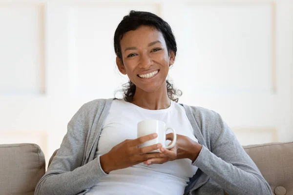Портрет счастливой расовой женщины расслабляется на диване и пьет кофе — стоковое фото