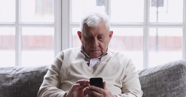 Улыбающийся дедушка 80-х с помощью мобильных телефонов сидит на диване — стоковое видео