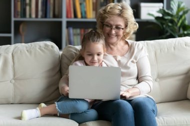 Smiling senior 60s granny use laptop with small granddaughter