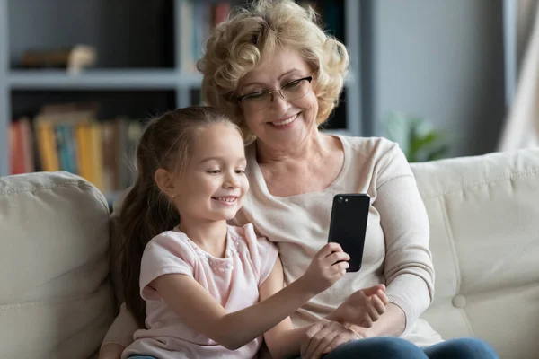 Улыбающаяся старшая бабушка и маленькая внучка делают селфи по телефону — стоковое фото