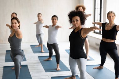 Grup halinde yoga dersinde Warrior Asana performansı sergileyen çok ırklı sporcular.