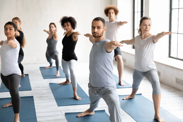 Семь начинающих мультиэтнических людей практикуют йогу, исполняя позу Воина II — стоковое фото