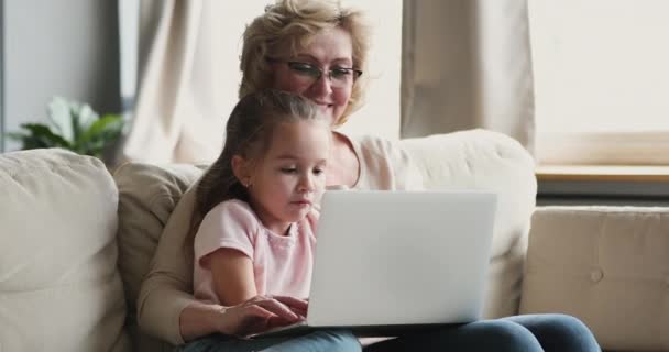 Lachende oudere oma en kleindochter met behulp van laptop samen op de bank — Stockvideo