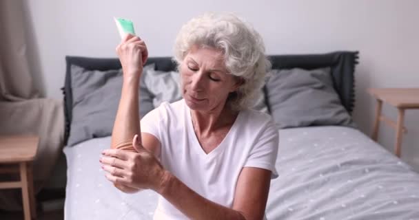 Mujer mayor sonriente aplicando crema en el codo sentada en la cama — Vídeo de stock