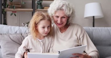 Senior granny reading book hugging granddaughter sitting on sofa